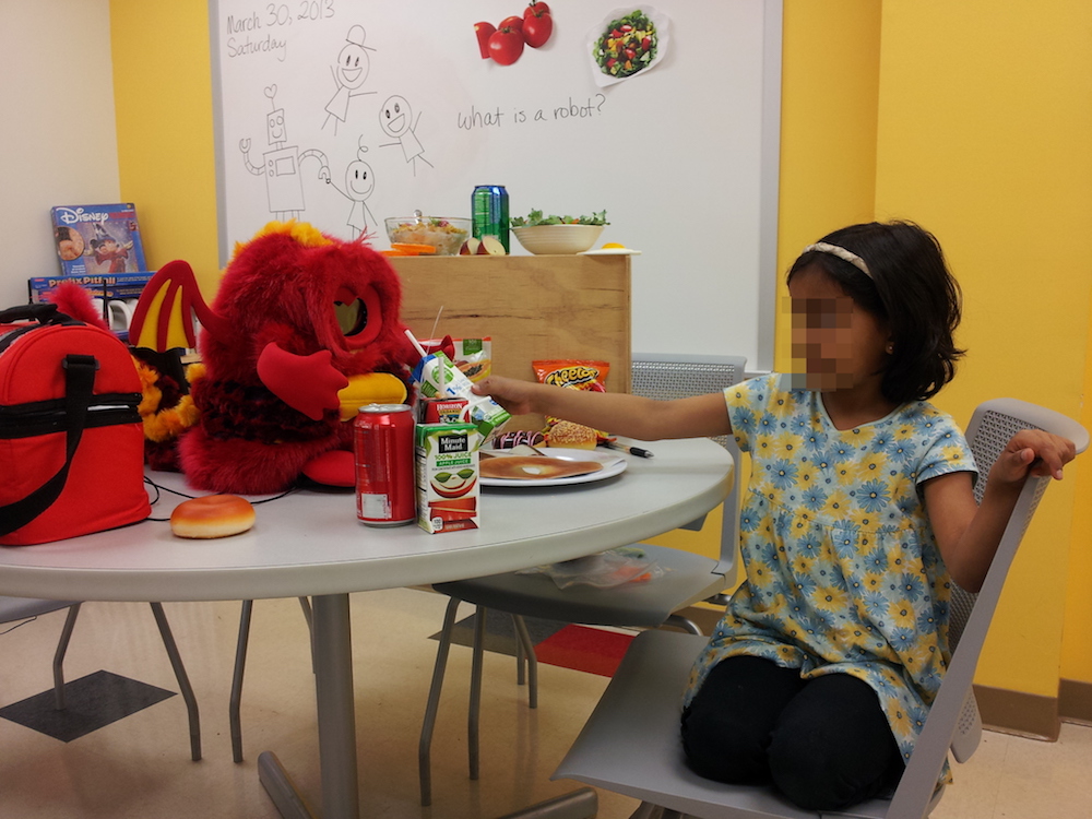 The small fuzzy robot Chili sits on a table among various food items, playing with a young girl of about five years of age.